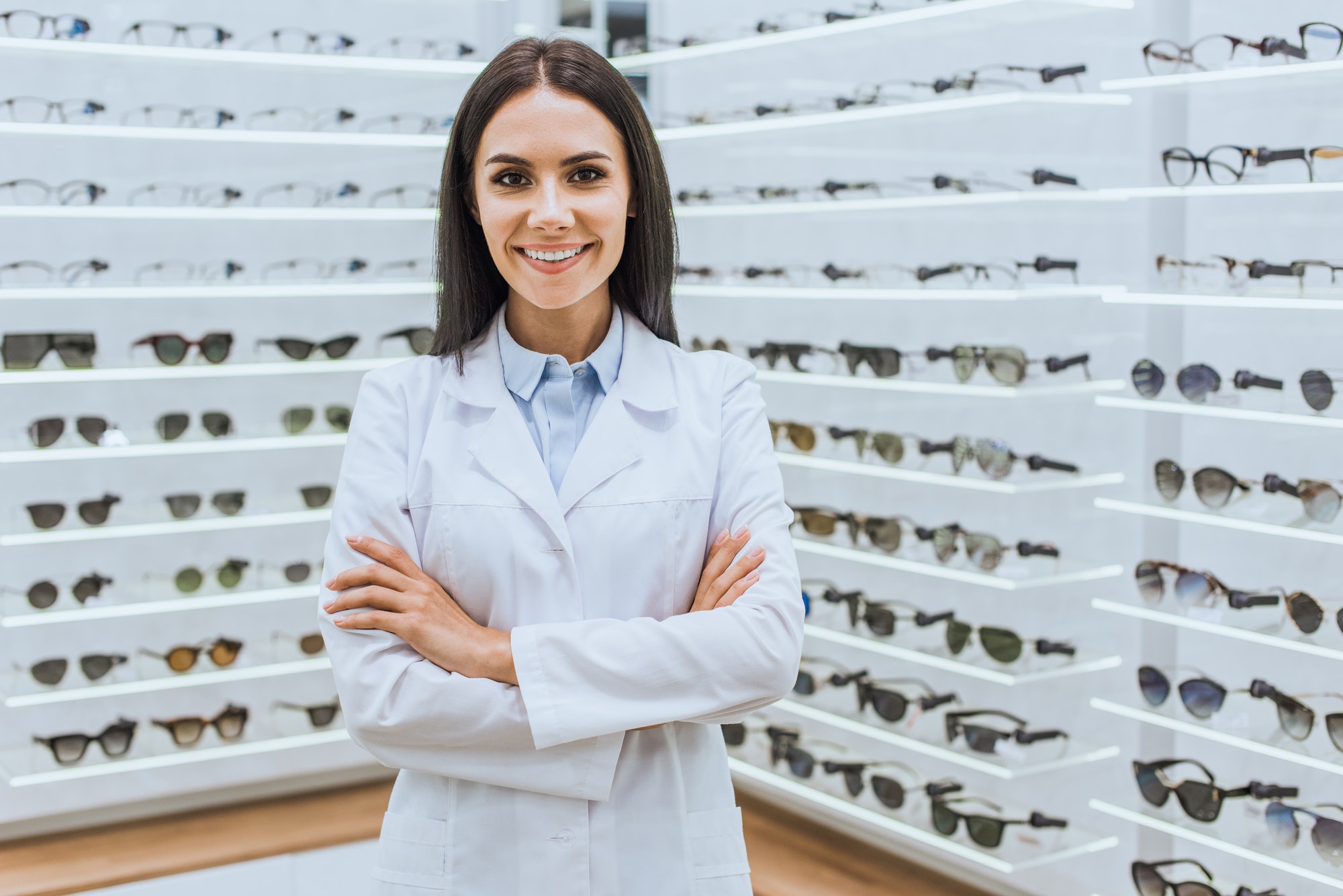 professional smiling optometrist posing with crossed arms near shelves with eyesight in optica